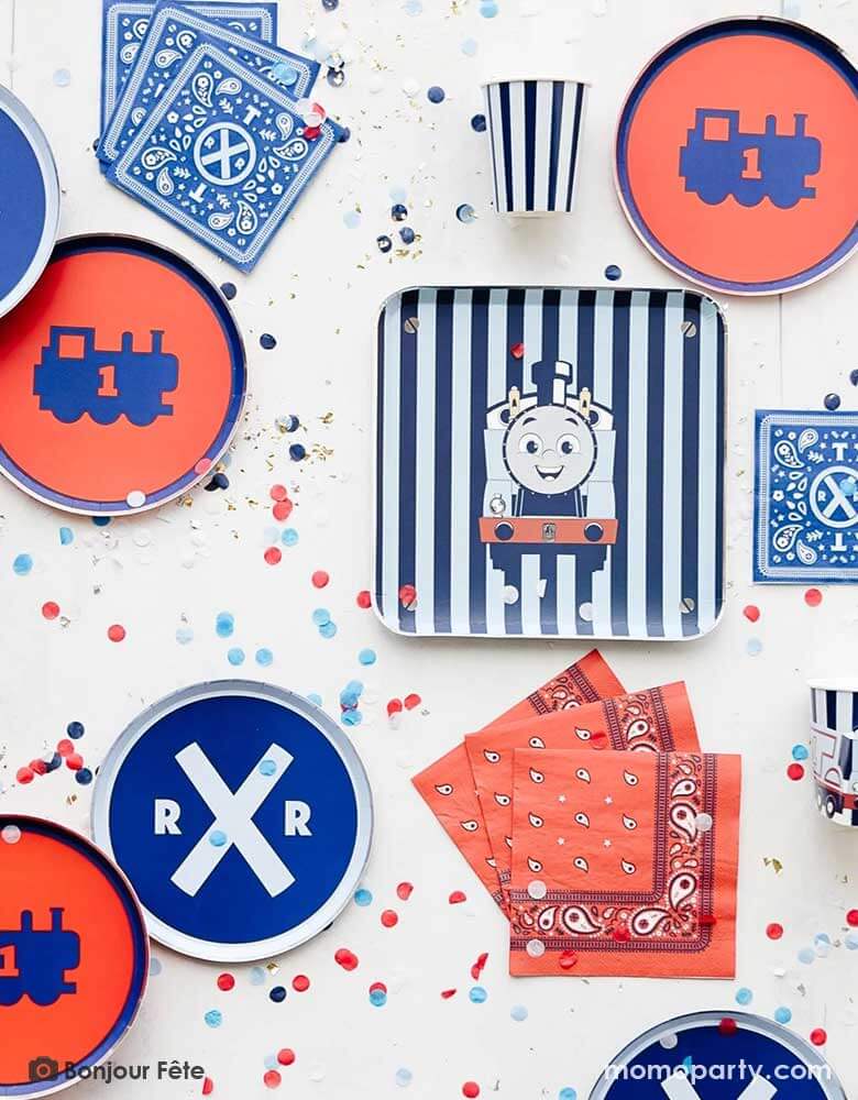 A table full of Thomas and Friend's themed party supplies from Momopartys by Bonjour Fete including blue striped square dinner plate with Thomas illustration on it, assorted side plates in blue and red, bandana inspired paper napkins in red and blue and Thomas 3D blue striped party cups, along with blue and red confetti spread out this is a perfect inspo for a modern kid's train themed Thomas and Friends birthday party!