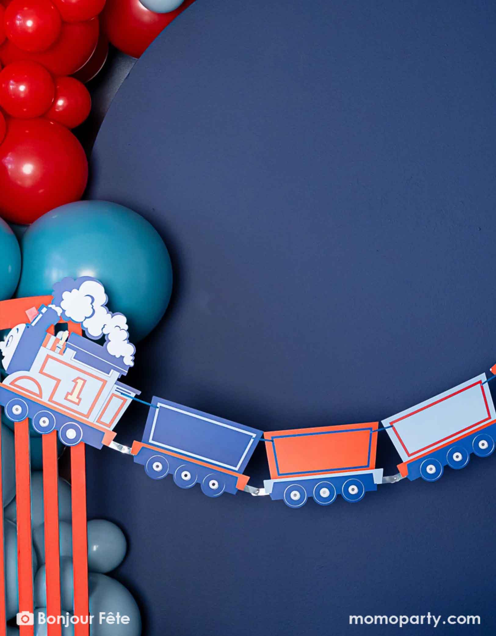 A close-up shot of Momopartys's Thomas the Train and Friends party garland hung on a blue wall which is decorated with a balloon garland in the classic train colors of blue and red.