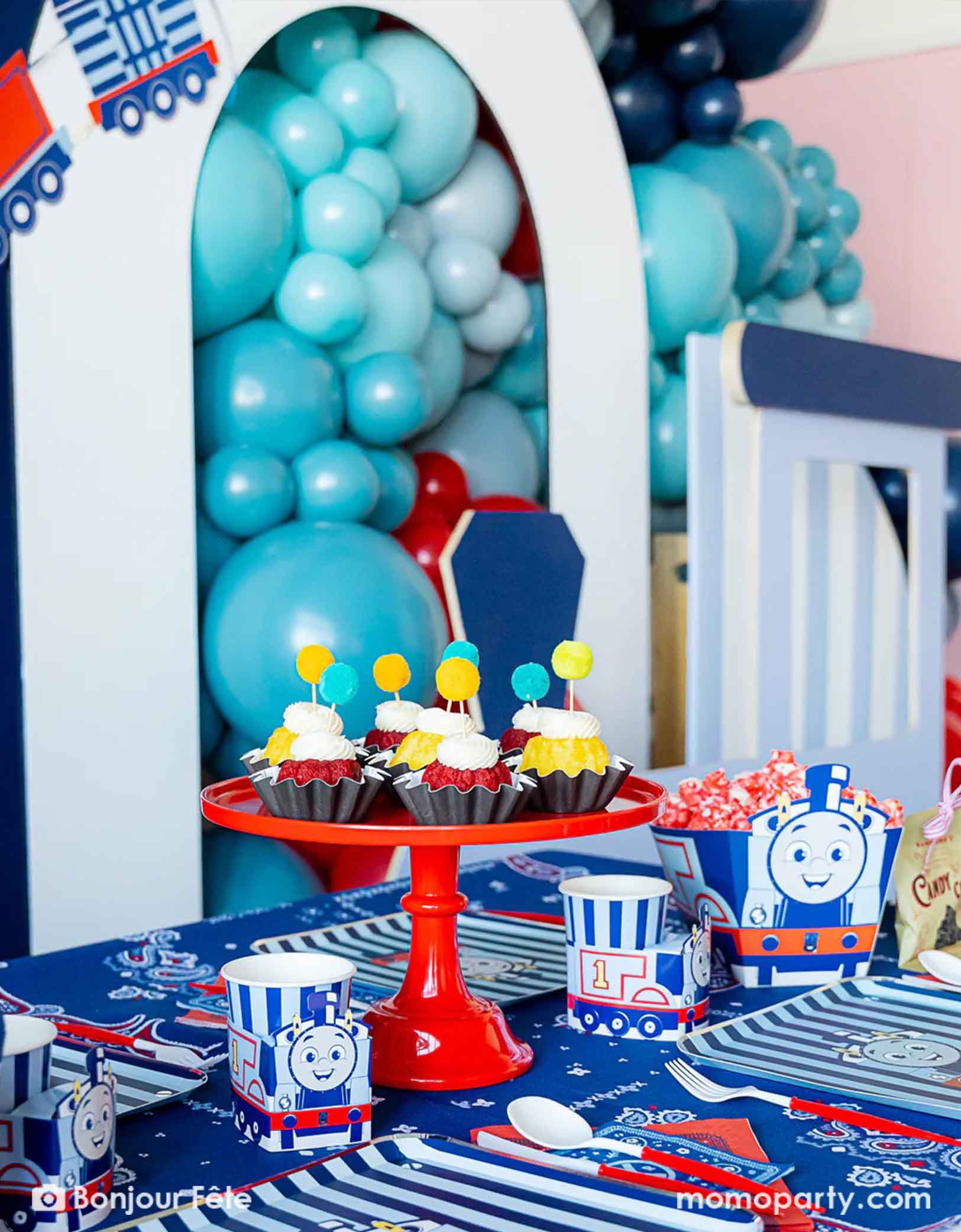 An adorable and festive Thomas the Train and Friends themed train party table featuring Momopartys's Thomas the Train themed party supplies including party tableware of plates, 3D cups, napkins treat boxes. The party table is covered with bandana inspired tablecloth to match the theme. In the middle of the table there's a red cake stand holding cupcakes topped with pom pom toppers. In the back is a blue & red balloon garland with a party garland, making this a perfect inspo for toddler's train party.