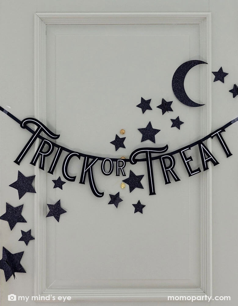 A gray front door decorated with Momopartys's 4ft trick or treat felt banner by My Mind's Eye. Around the felt banner are some black glittered stars and moon wall paper decorations, making this a great inspo for Halloween home decoration this spooky season!