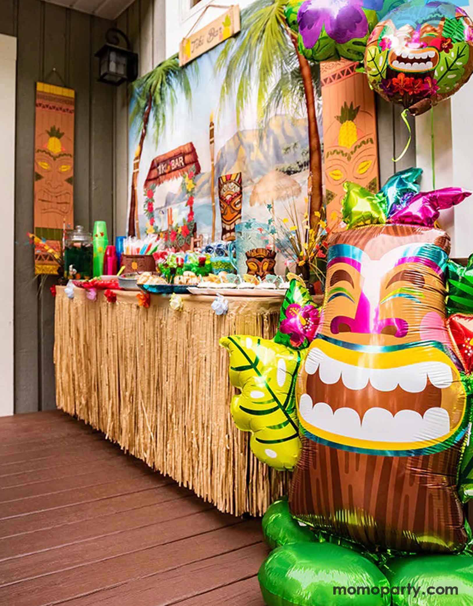 A festive tiki luau tropical themed party decoration set up featuring a tropical beach backdrop, Momopartys's colorful 53" airloonz tiki shaped foil balloon and a party table decorated with Hawaiian skirt around the table and various tropical themed treats and tableware. Perfect for a fun luau themed bash this summer!