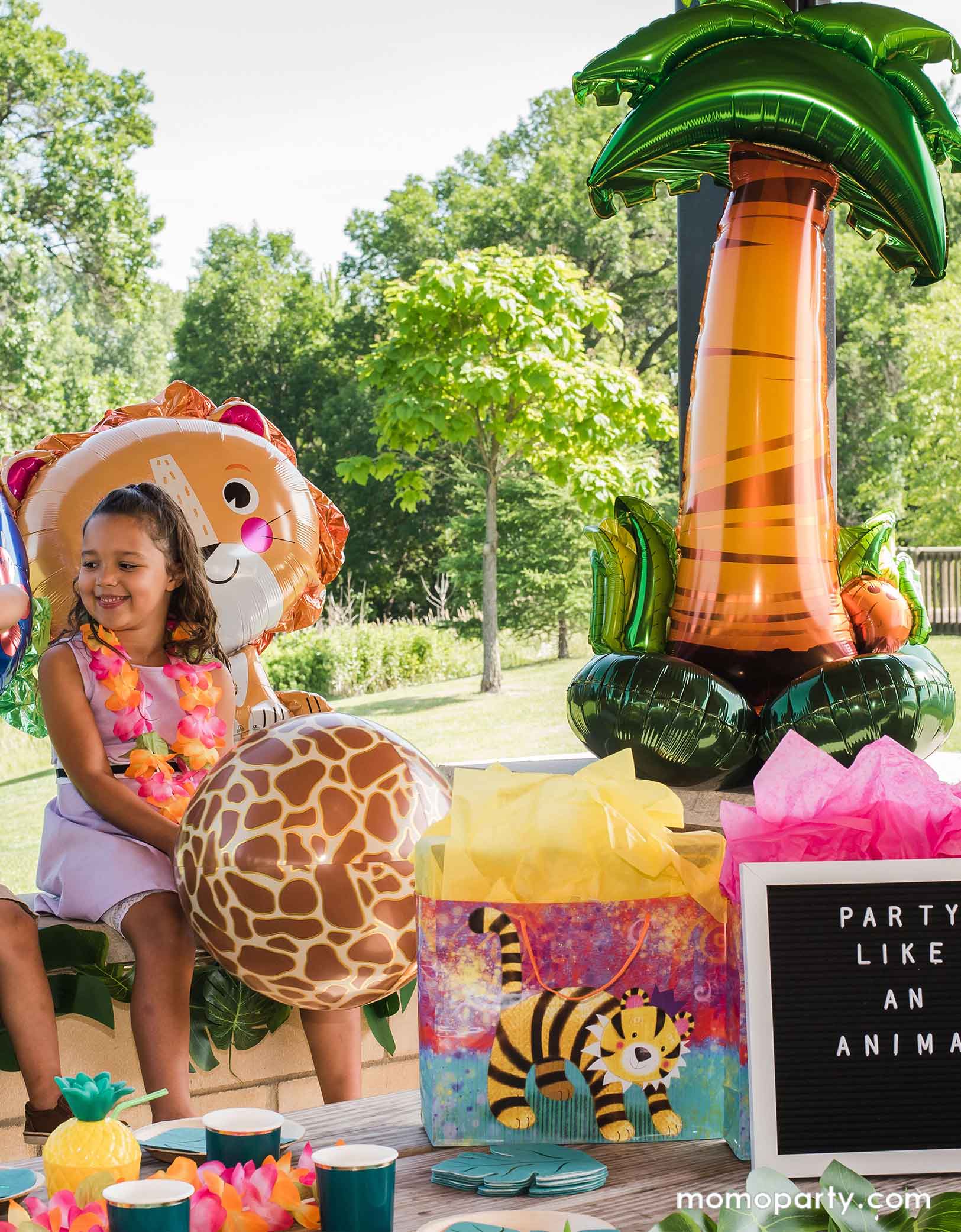 A little girl wearing Hawaiian leis stand next to Momopartys's 56" airloonz palm tree foil balloon in a tropical, luau, Hawaiian themed party decorated with animal themed foil balloons and palm tree party garland. On the party table there are some wild animal themed party favor bags and palm tree leaf shaped napkins and party cups.
