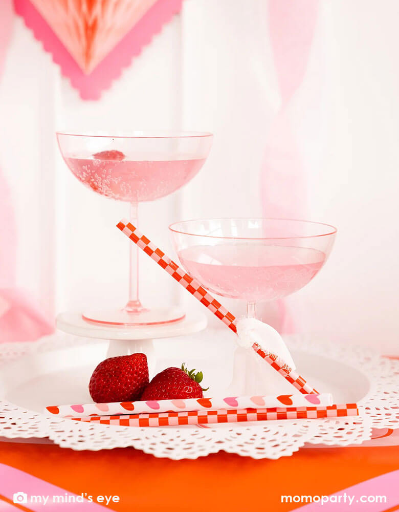 Two cocktail glasses with pink drink and a couple of strawberries on the side for a Valentine's Day celebration. Next to the drink there are Momopartys's heart you reusable straws with checkered and heart pattern on them by My Mind's Eye. In the back there's honeycomb heart shaped hanging decoration, making it a gorgeous inspiration for a sweet Valentine's Day party.