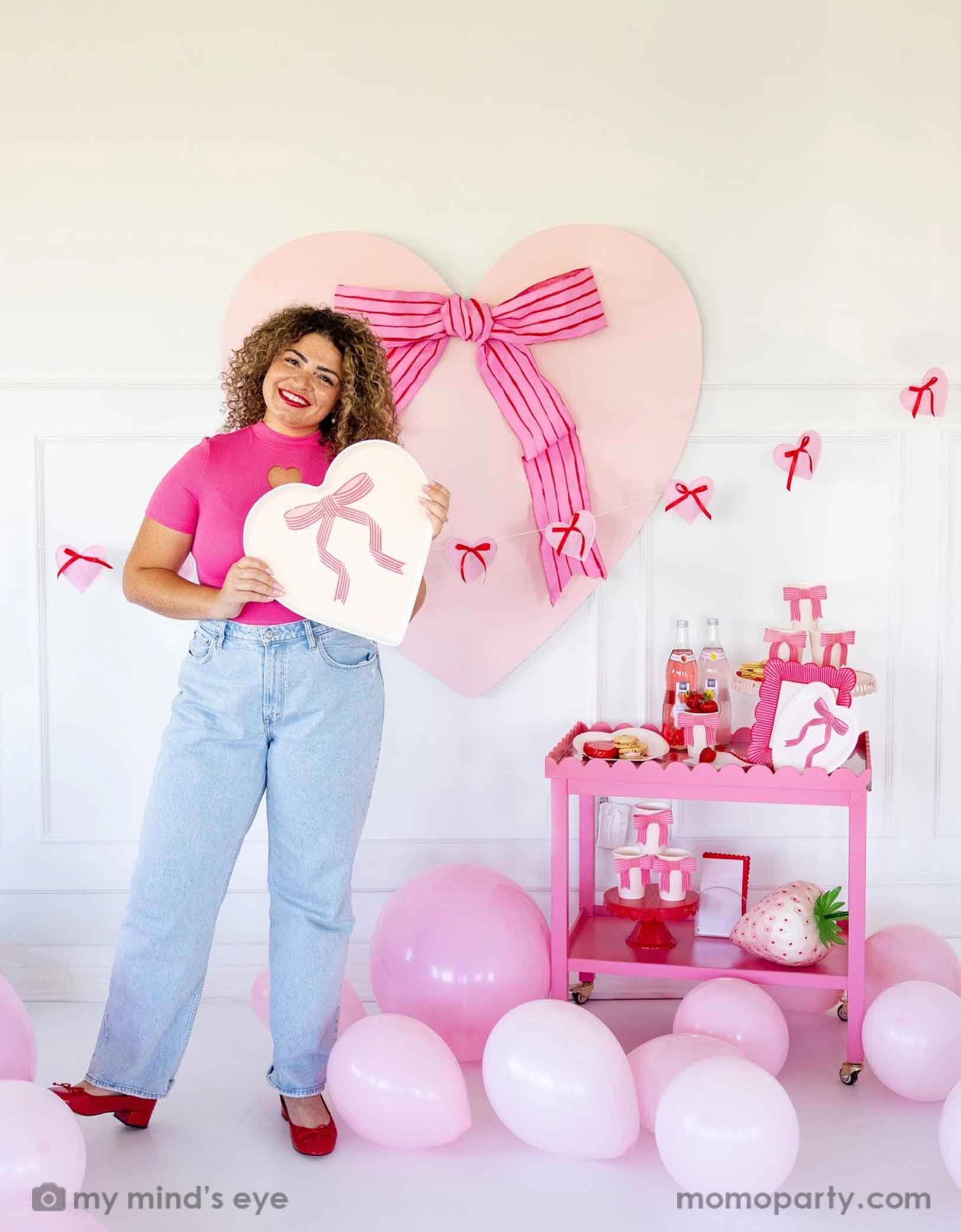 A lady with a hot pink top with red lipstick and red high heels holding Momopartys's 15" x 15" Bow Heart Tray by My Mind's Eye. She is standing next to a pink cart filled with Valentine's Day party supplies including plates, cups, napkins, along with drinks and snacks. In her back there's a giant pink heart shaped decoration with ribbon bow on the wall. With a floor of pink balloons, this makes a great scene for a sweet Valentine's Day celebration.