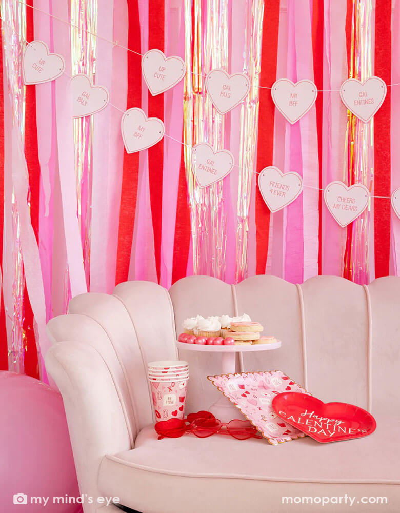 A pink room with the back wall decorated with red and pink streamers and a conversation heart candy banner. In the front there's a pink sofa featuring Momopartys's Valentine Scatter party cups and plates some heart shaped sunglasses and lots of Valentine inspired treats in red and pink.