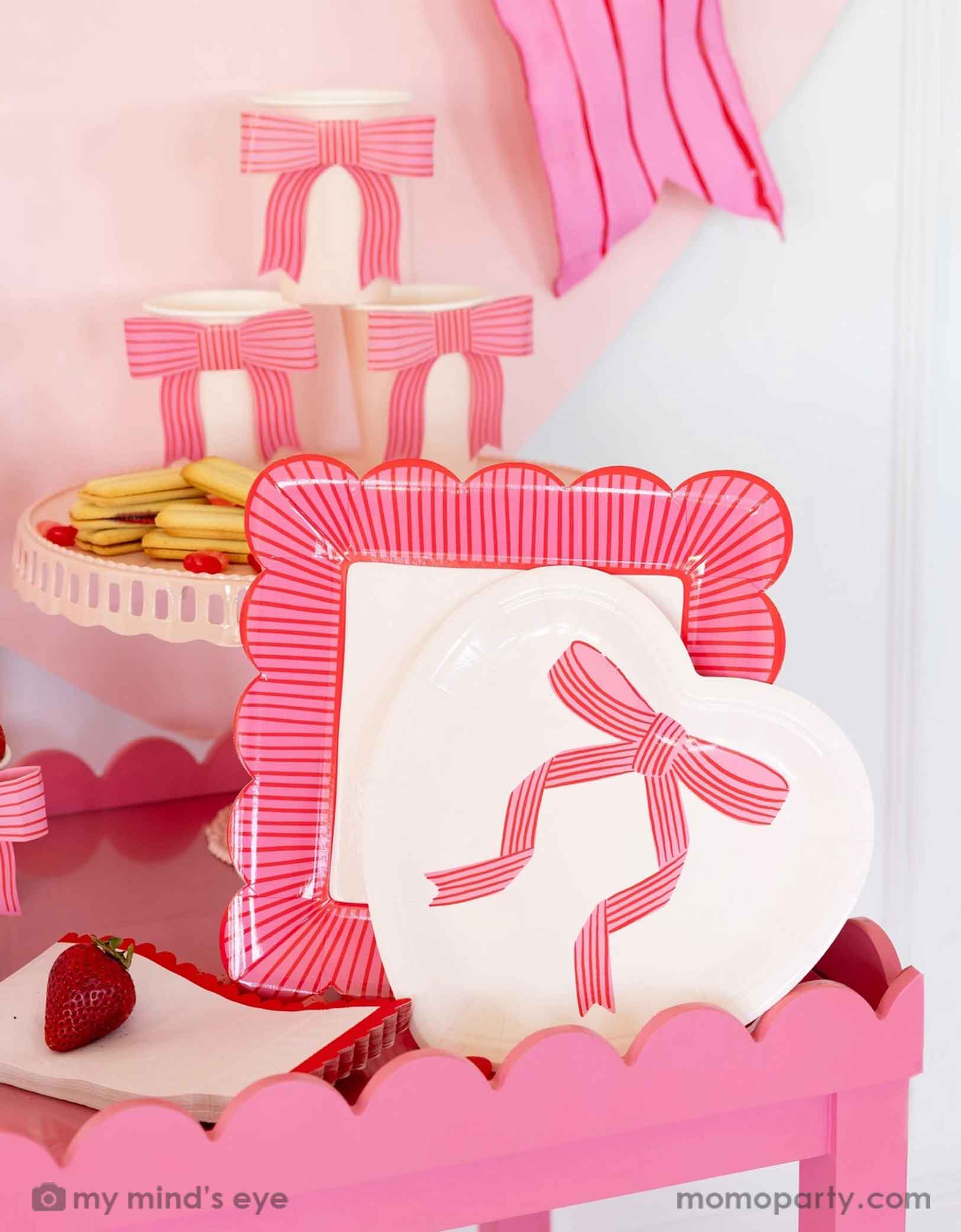 A Valentine's Day set up featuring Momopartys's bow heart Valentine's Day party collection including light pink heart shaped plate, bow party cups, striped scallop edged plate and scallop napkins. Around the tableware there are some heart shaped candies, cookies strawberries and some Valentine's Day themed snacks.