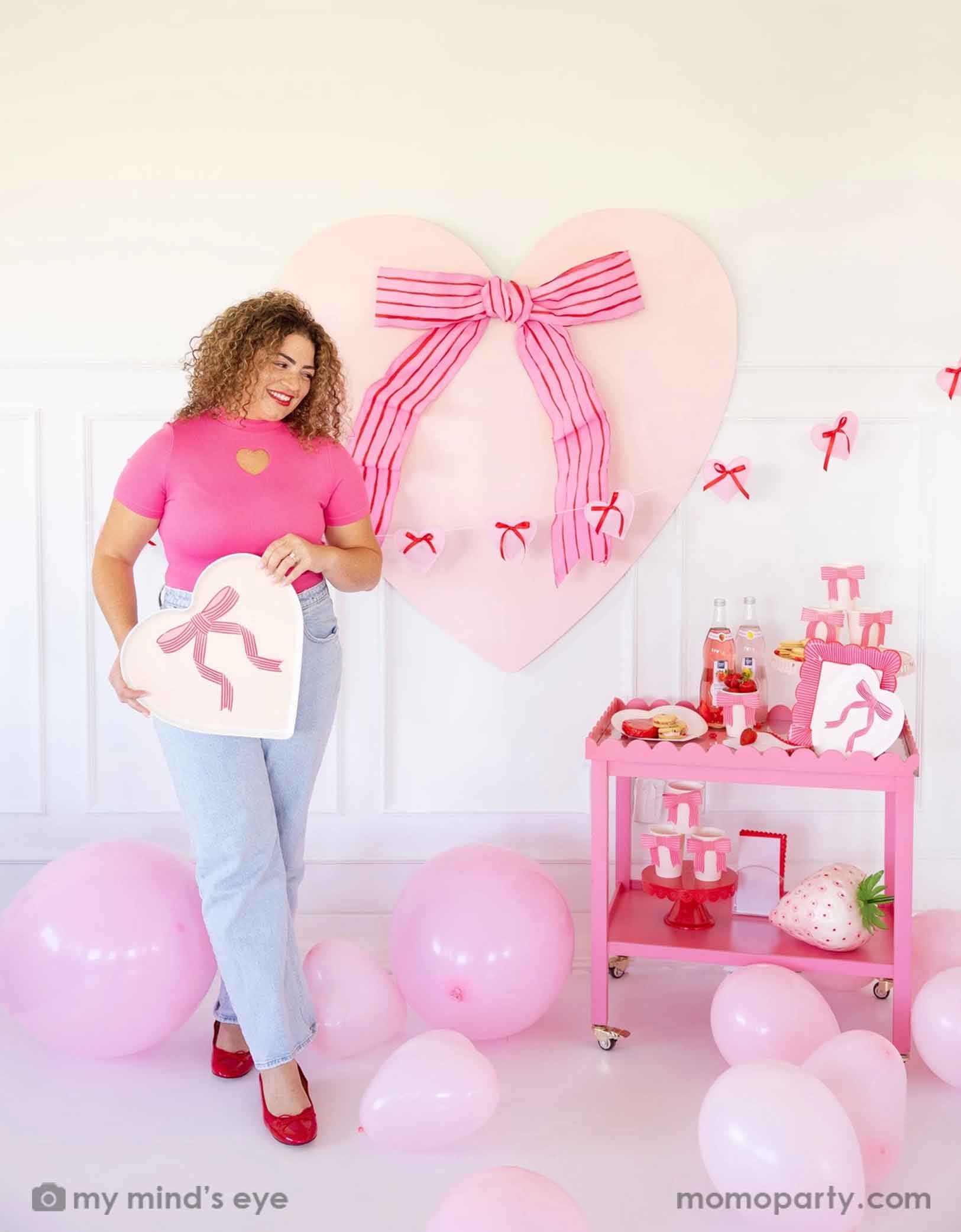 A lady with a hot pink top with red lipstick and red high heels holding Momopartys's 15" x 15" Bow Heart Tray by My Mind's Eye. She is standing next to a pink cart filled with Valentine's Day party supplies including plates, cups, napkins, along with drinks and snacks. In her back there's a giant pink heart shaped decoration with ribbon bow on the wall. With a floor of pink balloons, this makes a great scene for a sweet Valentine's Day celebration.