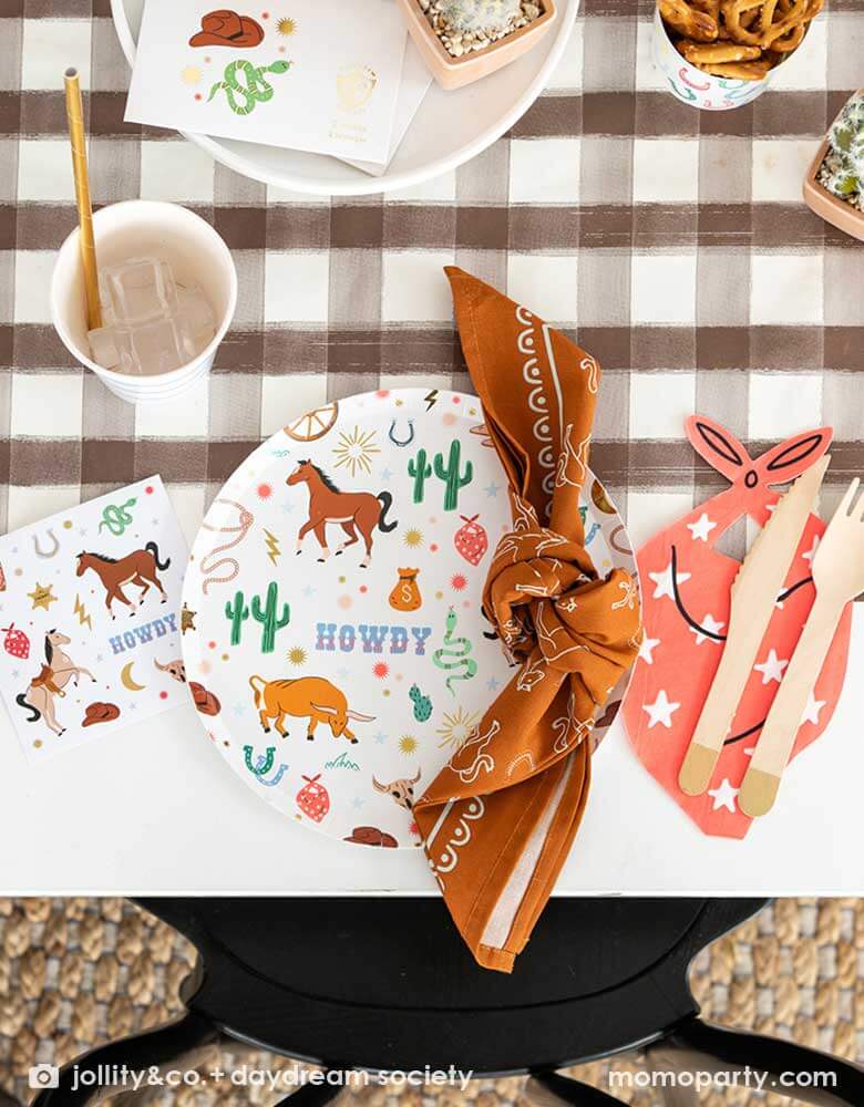 A western cowboy themed party table tablescape featuring Momopartys's cowboy themed tableware by Jollity Co. and Daydream Society including the round cowboy icon patterned round plate, the small napkin, the bandana shaped napkin, cowboy temporary tattoos all on a brown gingham table runner, making this a modern yet fun tablescape for kid's rodeo, Western or cowboy themed birthday celebration. A perfect inpso for kid's "First Rodeo" first birthday celebration.