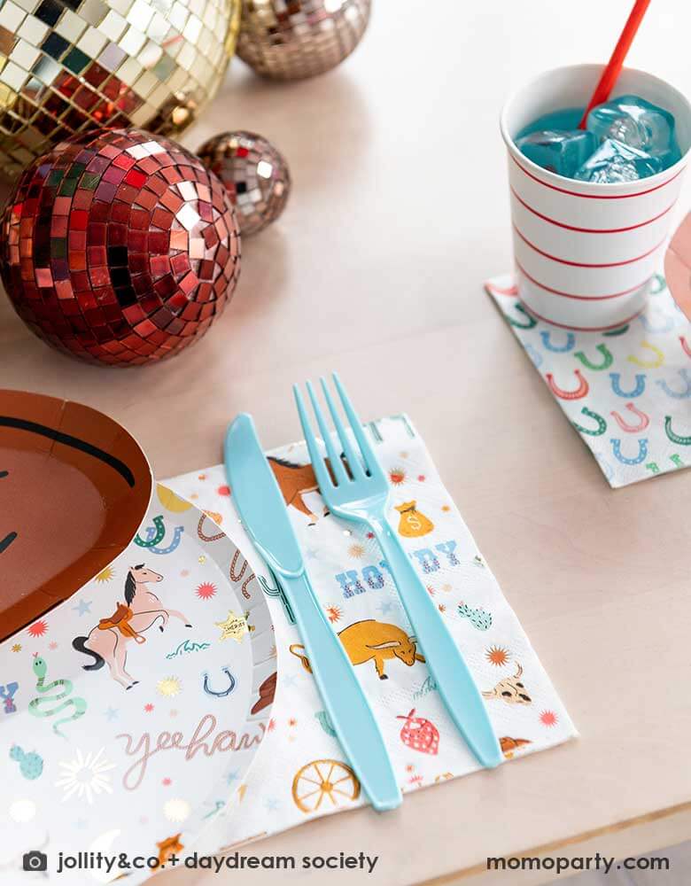 A closeup shot of a western cowboy themed party table tablescape featuring Momopartys's cowboy themed tableware by Jollity Co. and Daydream Society including the round cowboy icon patterned round plate, the large cowboy napkin, the small horseshoe napkin and cowboy hat shaped plate. With gold and red disco ball decorations on the table, making this a modern yet fun tablescape for kid's rodeo, Western or cowboy themed birthday celebration. A perfect inpso for kid's "First Rodeo" first birthday celebration.