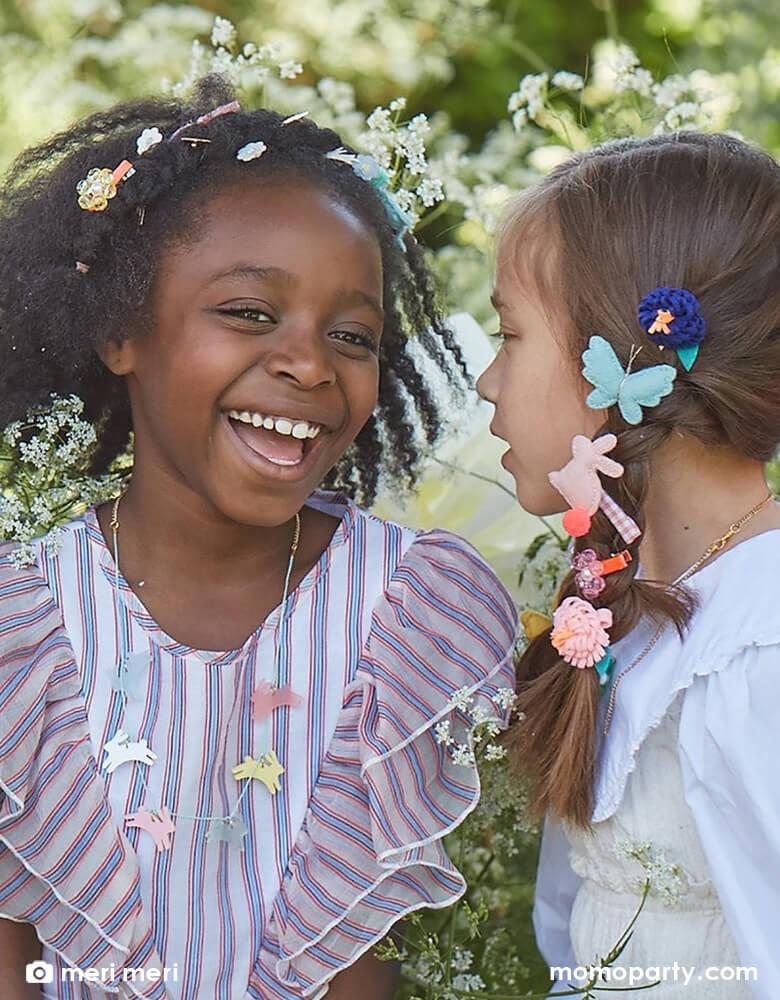 Two school aged girls chatting with each other wearing Meri Meri Easter themed accessories including bunny shaped felt hair clips, butterfly shaped felt hair chips and Meri Meri's acrylic leaping bunny necklace in 6 difference colors including frosted blue, white, frosted pink, frosted mint, yellow and pink, with a gold tone chain and mint cord necklace.
