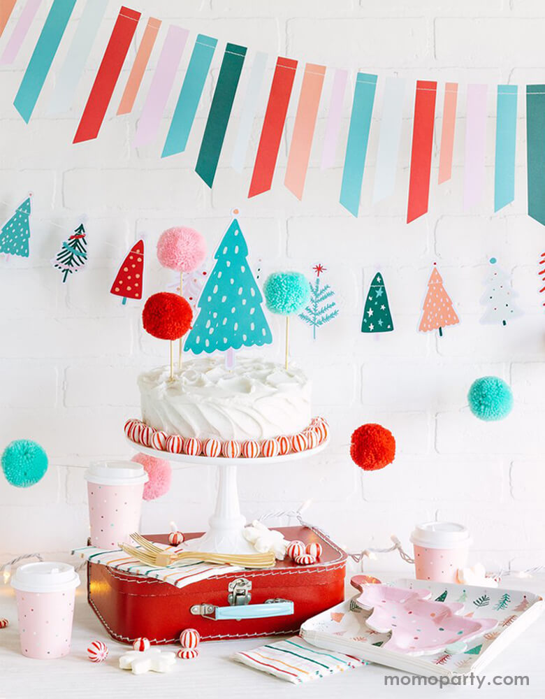 My Minds Eye & Oui Party Collab Christians party Dessert table set up with a buttercream white cake with pom pom cake topper and peppermint around, with a cake stand on top of a red vintage suitcase. a set of FA LA LA 9" FROSTING TREE PLATEs with FA LA LA CHRISTMAS 9" TREES PLATEs on the side, a 6ft Fa La La Christmas Tree Banner in cheery pastel colors and FA LA LA TICKER TAPE BANNER hanging on the wall as decoration for a pastel cute cozy christmas party