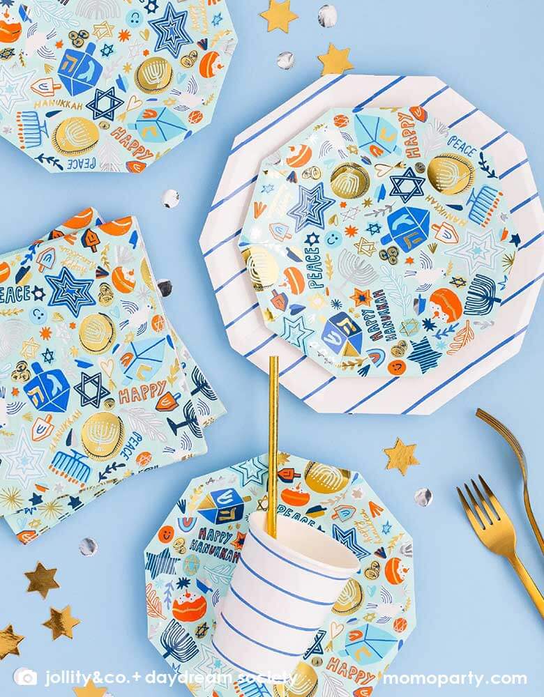 A tableware arrangement of Momopartys's 6.5" Festival of Lights Large Napkins and side plates by Daydream Society featuring Hanukkah themed symbol including dreidels, star of David, nine-branched candelabra, Menorah candles and Sufganiyah in blue colors with gold and silver foil detail, along with blue striped party cups and large plates, all together makes a modern Hanukkah tablescape