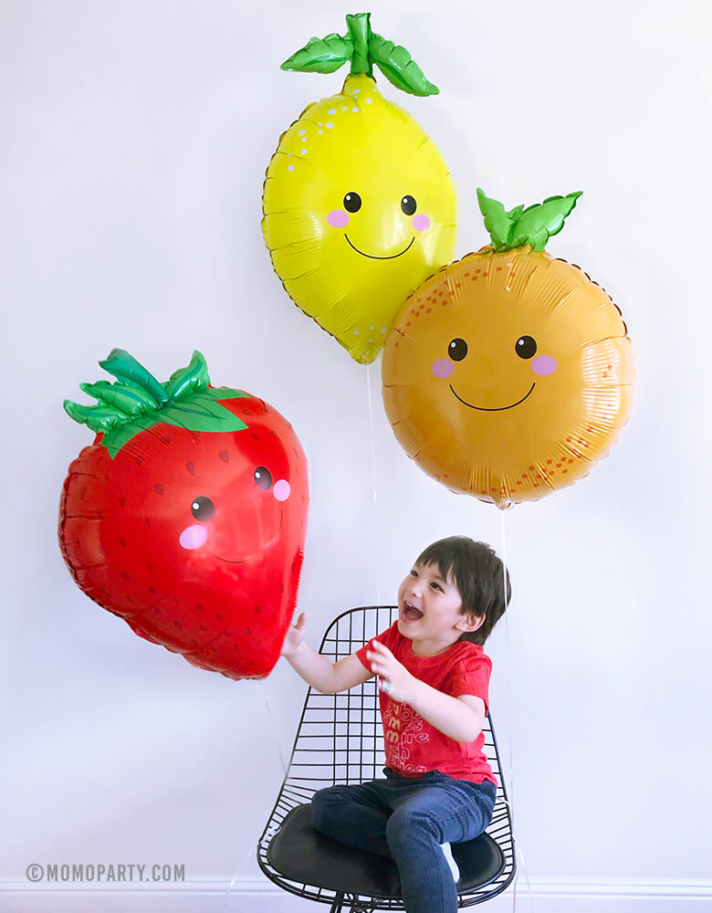 a happy boy catching 3 Betallic Produce Pal 29" Lemon, Strawberry and Orange Foil Mylar Balloons by sitting on a black Eames Wire Chair