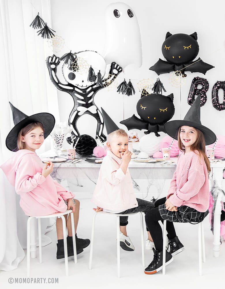 boy and girls wearing witch hats, seating in front of party table, celebrating a Pink Halloween. Room decorated with Party Deco Halloween Skeleton shaped Foil Mylar Balloon, Halloween Ghost Foil Mylar Balloon, Black bat foil balloon, and golden spiderwebs and black tassels garland. These halloween balloons also perfect for Kid-Friendly modern spooky halloween party, hocus pocus party trick-or-treating party, nightmare before christmas party, witch themed party and all halloween related celebrations