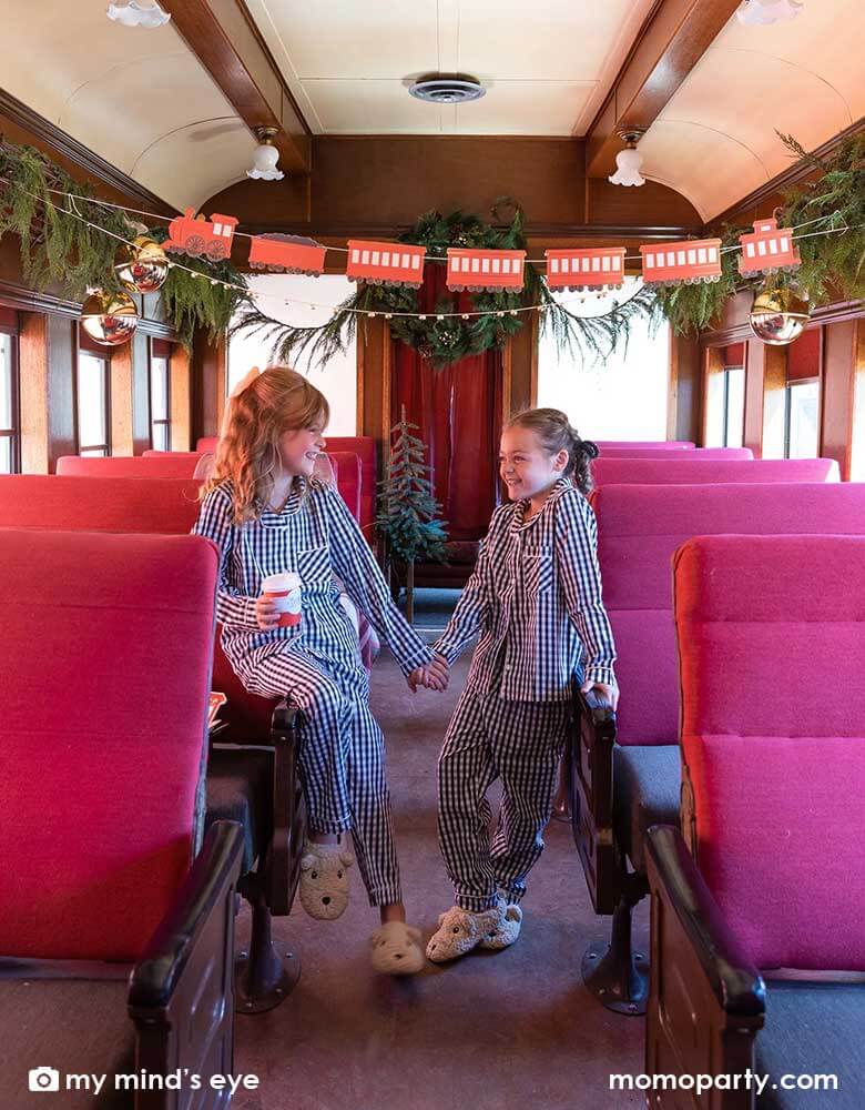 Two little girls in their Holiday pajamas holding North Pole Express red 8 oz to-go cups by My Mind's Eye sitting on a vintage Polar Express train cabin decorated with Momopartys's North Pole Express train and bell garland set, a perfect idea for this Holiday season's celebration with family and kids.