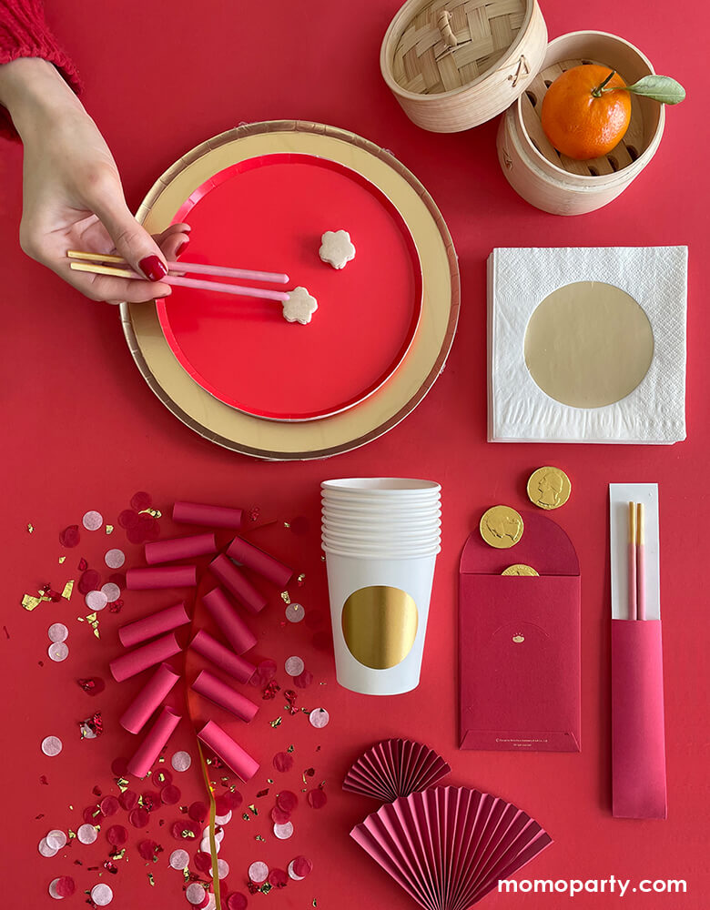 Momo Party's Modern Lunar-New-Year Party ideas with red small round plate overlap a GOLD LARGE PLATES, with a hand holding 2 strawberry flavor pocky sticks as chopstick, a gold dot cup, a red envelope with gold chocolate, gold dot napkin, a mandarin inside a mini Bamboo Steamer, paper rolls with confetti as firework. With all the gold and red that symbolizes happiness and prosperity over a red background, These modern and fun party supplies is new way to celebrate Lunar New Year!