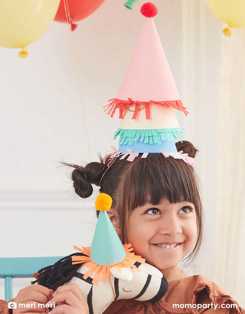 A girl wearing multiple party hats in bright colors holding a zebra stuffy what wears Momopartys's 2.25 x 4" mini party hat by Meri Meri.