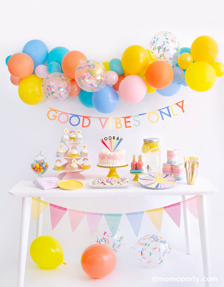 Momopartys Good Vibes Only Party Box Kit. Included Meri Meri Rainbow Sun Dinner Plates, Oh Happy Day Yellow Side Plates, Meri Meri Rainbow Sun Fringe Cups, Party Deco Good Vibes Napkins, Yellow Wooden Cutlery, Mixed Pastel Striped Straws, cupcake with rainbow and daisy candles. Meri Meri Hooray cake topper on a dripping cake, colorful gumballs, Lemonade on the table. Meri Meri Multicolor Bunting Banner spelled as "good vibes only" text , Good Vibes 6-foot- Balloon Garland Cloud as decoration.