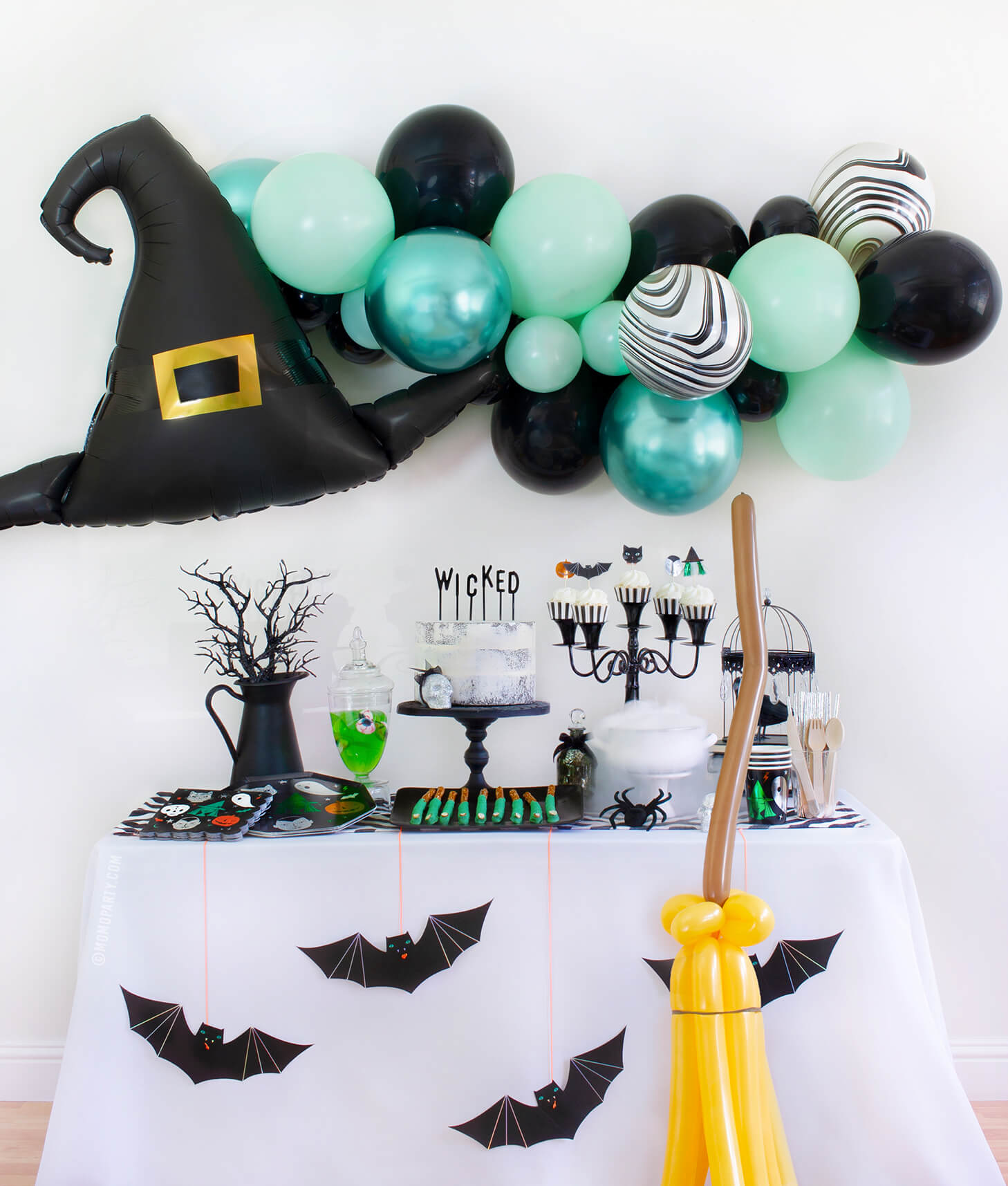 Momopartys 2020 Witch Please themed Halloween Party at home inspiration look with Anagram Witch Hat Satin Foil Balloon and pastel Green toned Latex Balloon Garland for wall decoration. Meri Meri Halloween Motif Dinner Plates, napkins and cups, cake with letter board cake topper spelled of "Wicked", witch cauldron, Apple jelly with eyeball candy in a jar, witch fingers pretzels on the dessert table, Meri Meri Hanging Bats and balloon animal witch broomstick in front of the table