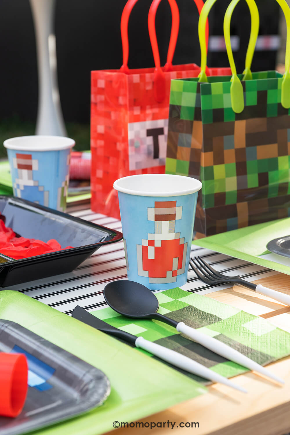 Minecraft themed Birthday Party Ideas by Momopartys. Featuring blue Minecraft potion paper cup with image of a potion bottle print, Minecraft gray side plate and modern sophisticated large lime green dinner plates, beautiful black and white cutlery sets, an pixel TNT theme goodie bags, all on the black striped table runner of the kids picnic table.