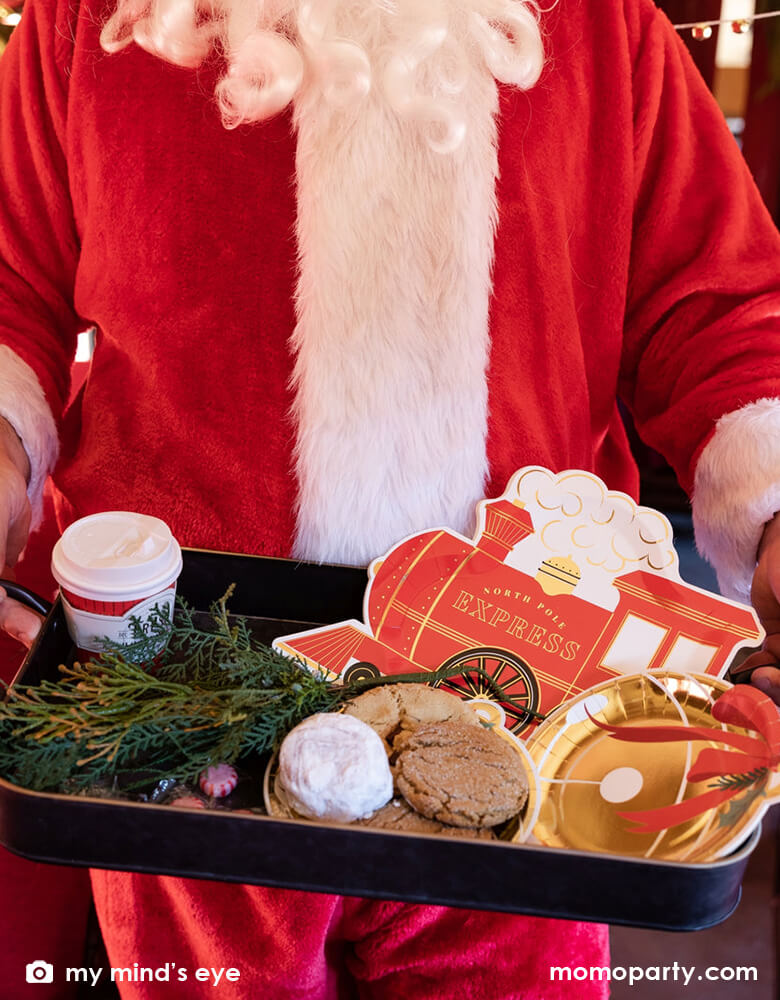 Santa holding a vintage tin tray filled with holly branches, holiday treats and cookies and Momopartys's North Pole Express train shaped plates, bell shaped side plates and polar express red 8 oz to-go cups by My Mind's Eye on a vintage Polar Express train cabin red leather seat, ready for a festive Holiday celebration for family and kids.