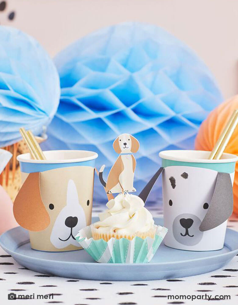 A party table featuring adorable dog themed party goods including Momopartys's 9oz puppy party cups by Meri Meri and cupcakes decorated with Meri Meri's puppy cupcake toppers. In the back it's a bunch of blue and peach colored honeycomb decorations, all together makes a great inspiration for a modern and chic dog themed party for kids or dog-loving adults.