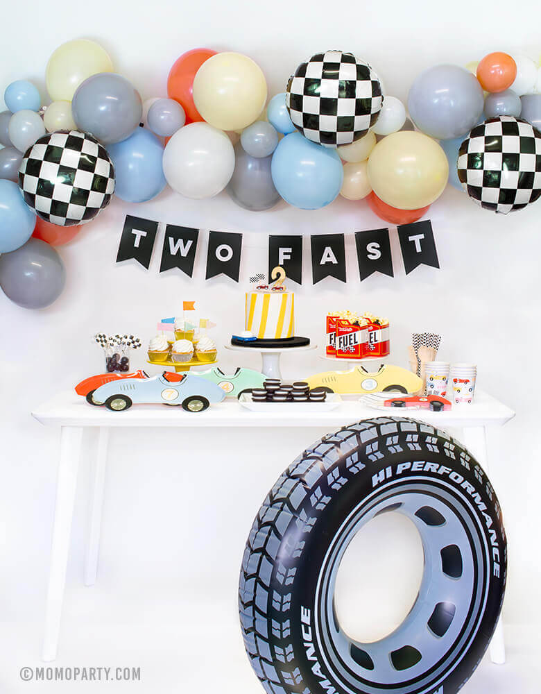 Momopartys Race Car Box, set up inspiration with Meri Meri race car paper plates, race car shape napkins, race car graphic party paper cups, black strip small plate, neon confetti flag cupcake kit, race Car candles and gold #2 candle over a cake, Fuel treat favor box with popcorns as tableware, colorful balloon cloud, black costume letter banner with script of "Two Fast", checkerboard foil balloon and Inflatable giant tire tube as decorations, for a 2 years old modern Race car, car themed birthday party
