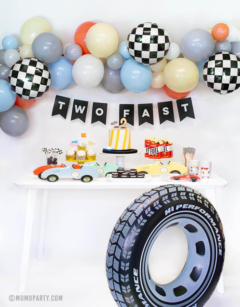 Momopartys Race Car Box, set up inspiration with Meri Meri race car paper plates, race car shape napkins, race car graphic party paper cups, black strip small plate, neon confetti flag cupcake kit, race Car candles and gold #2 candle over a cake, Fuel treat favor box with popcorns as tableware, colorful balloon cloud, black costume letter banner with script of "Two Fast", checkerboard foil balloon and Inflatable giant tire tube as decorations, for a 2 years old modern Race car, car themed birthday party