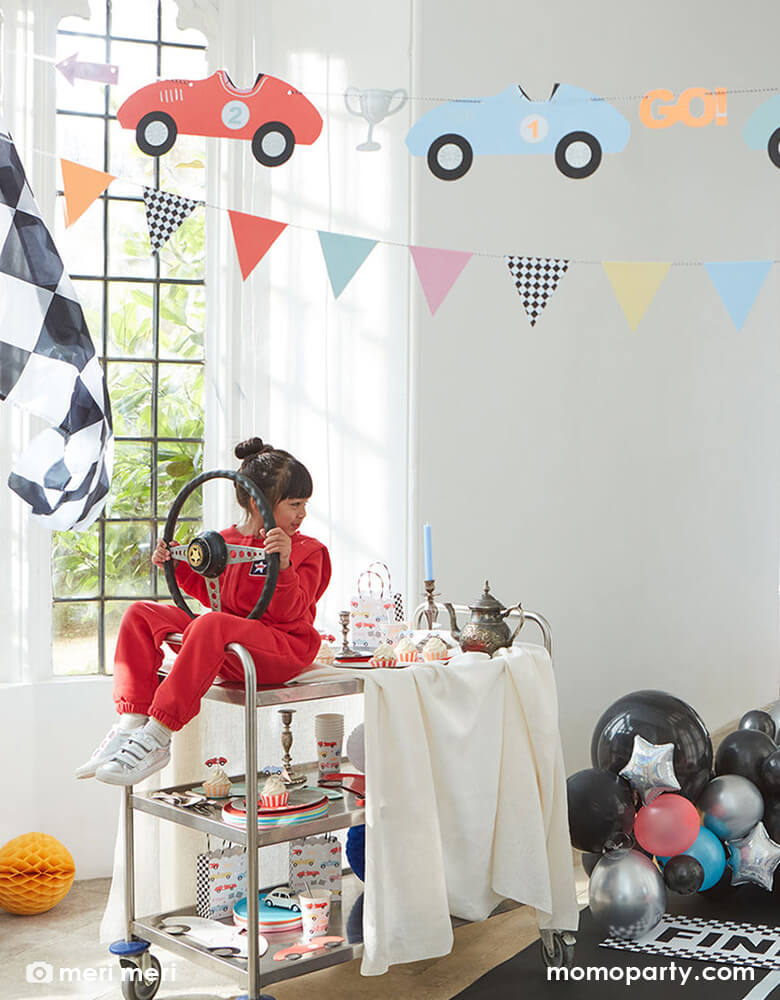 Race car party in the house, decorated with Meri Meri Race Cars Garland, balloons, race car checker flags, with a birthday girl wearing a red racing suit, holding a car Steering Wheel pretending like driving by sitting on a cart with all the car themed party tableware