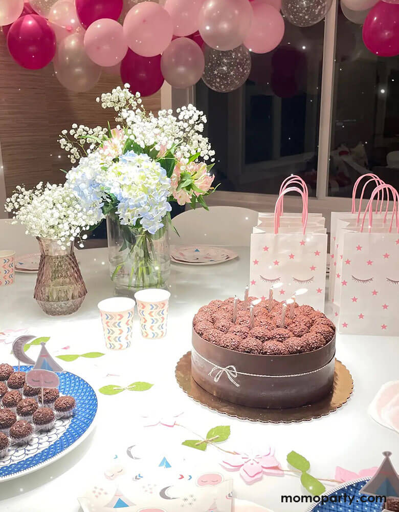 A sleepover slumber party featuring a chocolate cake, some cupcakes and Momopartys's 8.2 x 5.9 x 3.15 inches sleepover party bags with sleepy eyes and pink star designs by Pooka Party, comes in set of 8. In the background the room was decorated with pink, rose, and rose gold balloon garland with beautiful flower bouquet, makes a great inspiration for girls birthday party ideas.