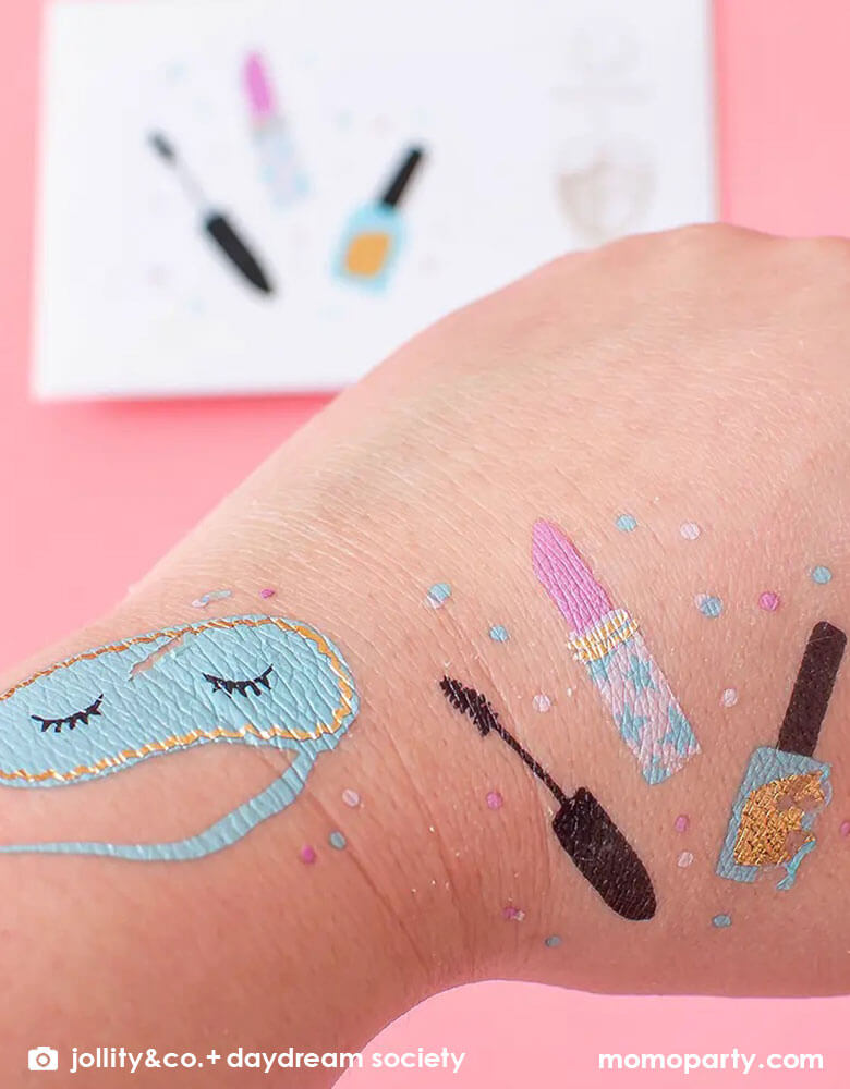 A close up of a girl's hand covered with Momopartys's Sweet Dreams Slumber party temporary tattoos by Daydream Society, comes in a set of 2 temporary tattoos, on in a design of an adorable eye mask, the other with nail polish bottle, lip stick and eye lash mascara, this set makes great party activity for girl's sleepover pajama party.