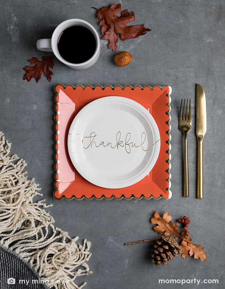 Table top view of My mind's Eye 7 inches Thankful Grateful Plates with "thankful" printed in a beautiful script in gold foil on the cream paper plates, layered with Orange with golden foil scalloped large plates, there are golden cutlery, a cup of coffee, some dry Maple leave, nuts around on top of the grey quartz countertop. These modern party supplies are Perfect for your Thanksgiving celebration dessert or appetizers.
