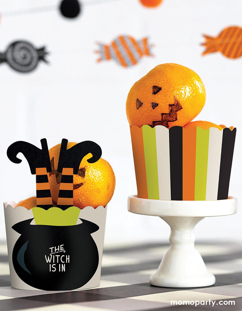 Table top with tangerines in the My Minds Eye - WITCH IS IN FOOD CUPS. one with Witch Leg Stakes with "the witch is in" text in the Cauldron. another cups in a mint, orange, white and black stripe design with tangerine drawed as a jack-o-lantern. These fun halloween themed baking cups are perfect for baking Halloween cupcakes right in the oven.