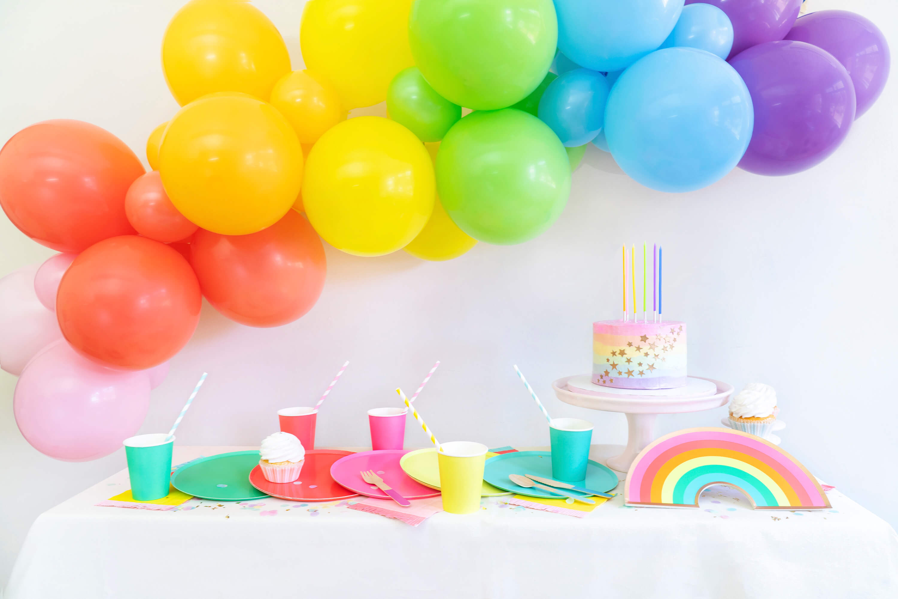 Momopartys Modern rainbow party table look with colorful rainbow large paper plates, cups, pastel rainbow cake with rainbow candles, rainbow balloon garland with pink, coral, orange, yellow, lime, blue and purple latex balloon