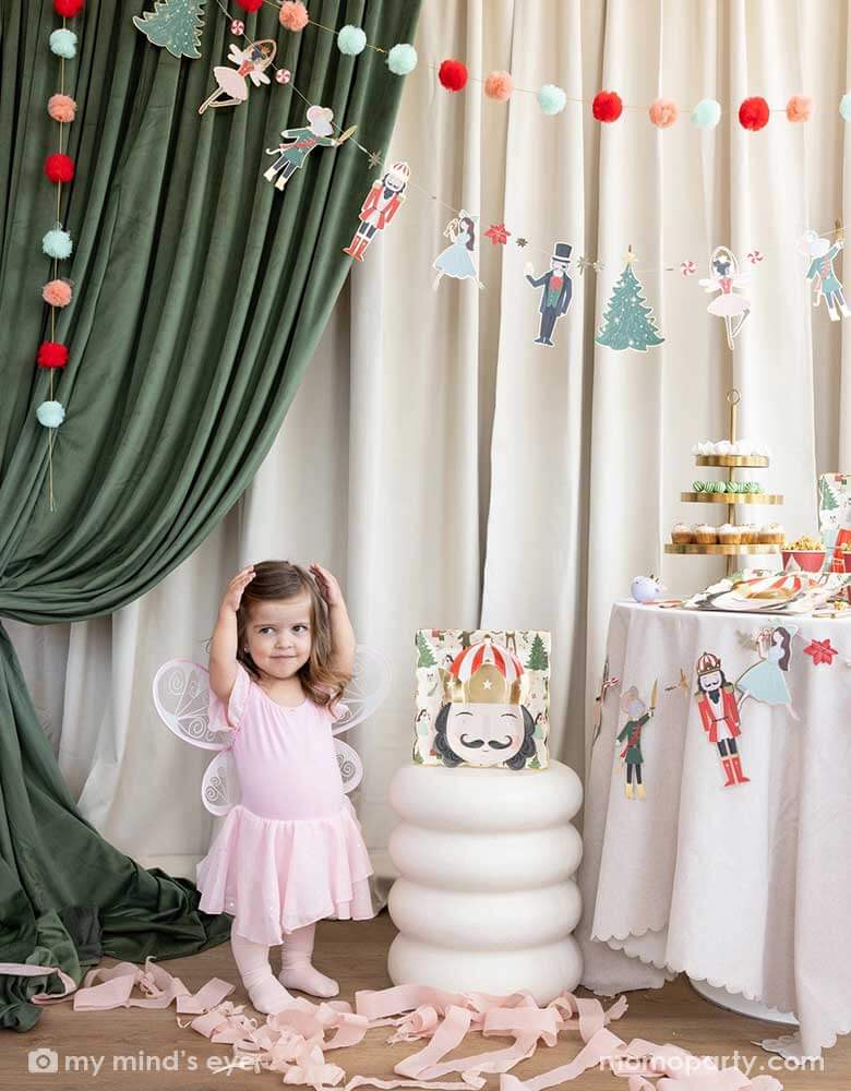 A little girl doing a ballet pose standing next to a Holiday party table filled with Holiday treats and Nutcracker themed party supplies including party plates, cups, party garland. In her back there's a stage curtain in satin green and beige which is decorated with Momopartys's Nutcracker garland with pom-poms which adds a festive touch to the Holiday decoration.