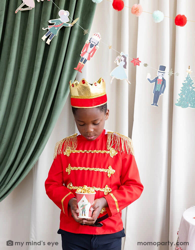 A boy dressed up as Nutcracker's soldier holding Momopartys's 12 oz Nutcracker party cup filled with popcorn. In his back there's a stage curtain in satin green and beige which is decorated with Momopartys's Nutcracker garland with pom-poms which adds a festive touch to the Holiday decoration.