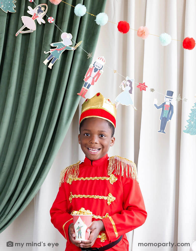 A boy dressed up as Nutcracker's soldier holding Momopartys's 12 oz Nutcracker party cup filled with popcorn. In his back there's a stage curtain in satin green and beige which is decorated with Momopartys's Nutcracker garland with pom-poms which adds a festive touch to the Holiday decoration.
