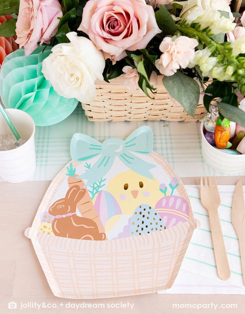 An adorable kid's friendly Easter table setting features Momopartys's Easter Fun basket shaped plates, mint striped plates, party cups by Daydream Society. In the middle there's light blue gingham table runner, Easter Egg tissue honeycomb in mint and peach and a beautiful Easter flower arrangement in a basket. On the table there's also a small treat cup filled with Easter candies and chocolates, making this an adorable Easter tablescape.