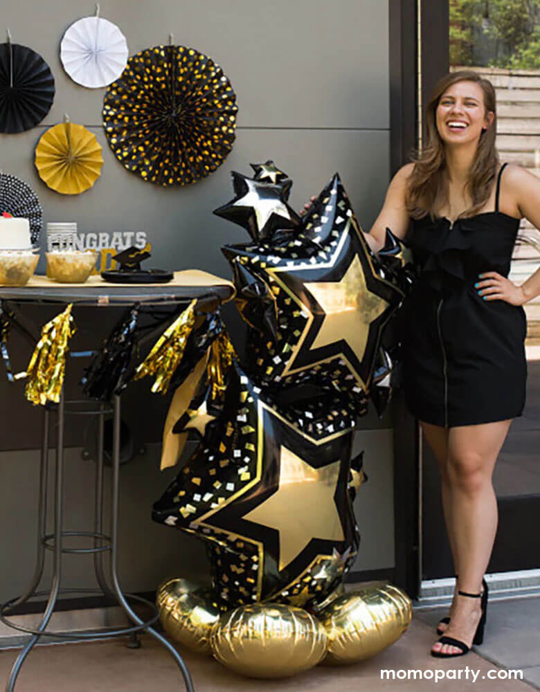 A collage or high school graduate standing next to Momopartys's 59" Airloonz Black and Gold Star Cluster Foil Balloon by Anagram Balloons next to a dessert table decorated with graduation themed decorations including black and gold party fans, tassels, "Congrats" candles to celebrate her graduation day.