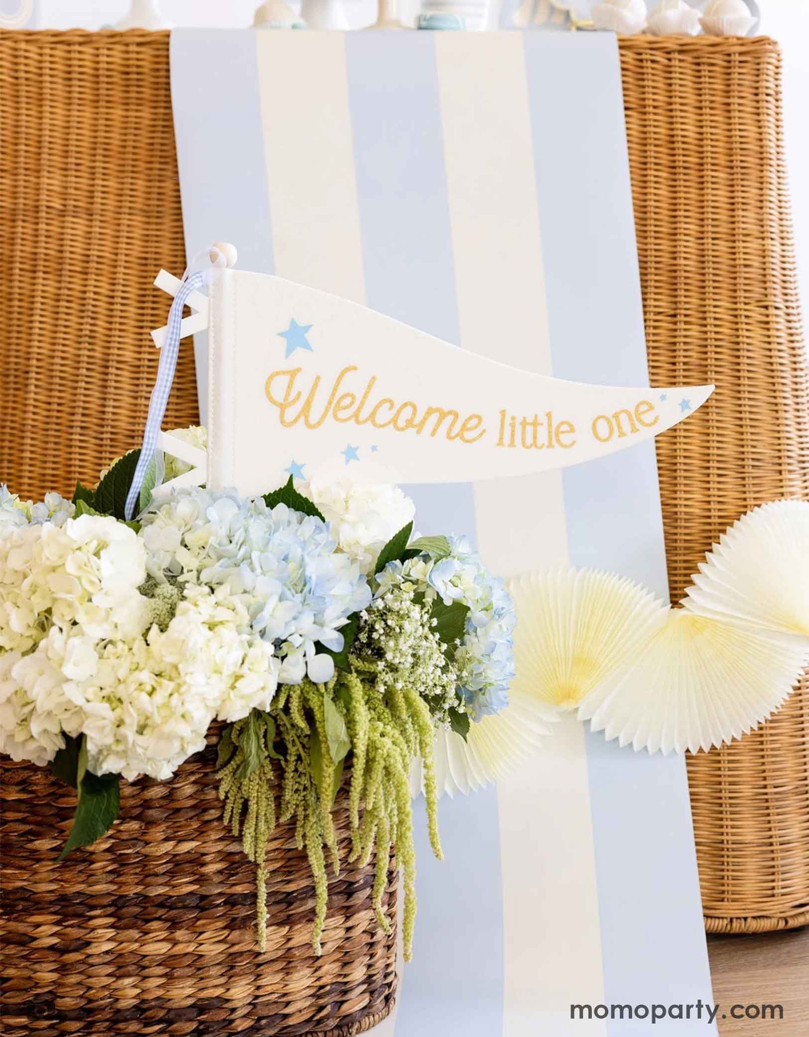 A modern and clean boy baby shower set up featuring straw-strung table covered with Momopartys's baby blue and white striped table runner with flowers in a basket and 'Welcome little one' sign at the bottom.