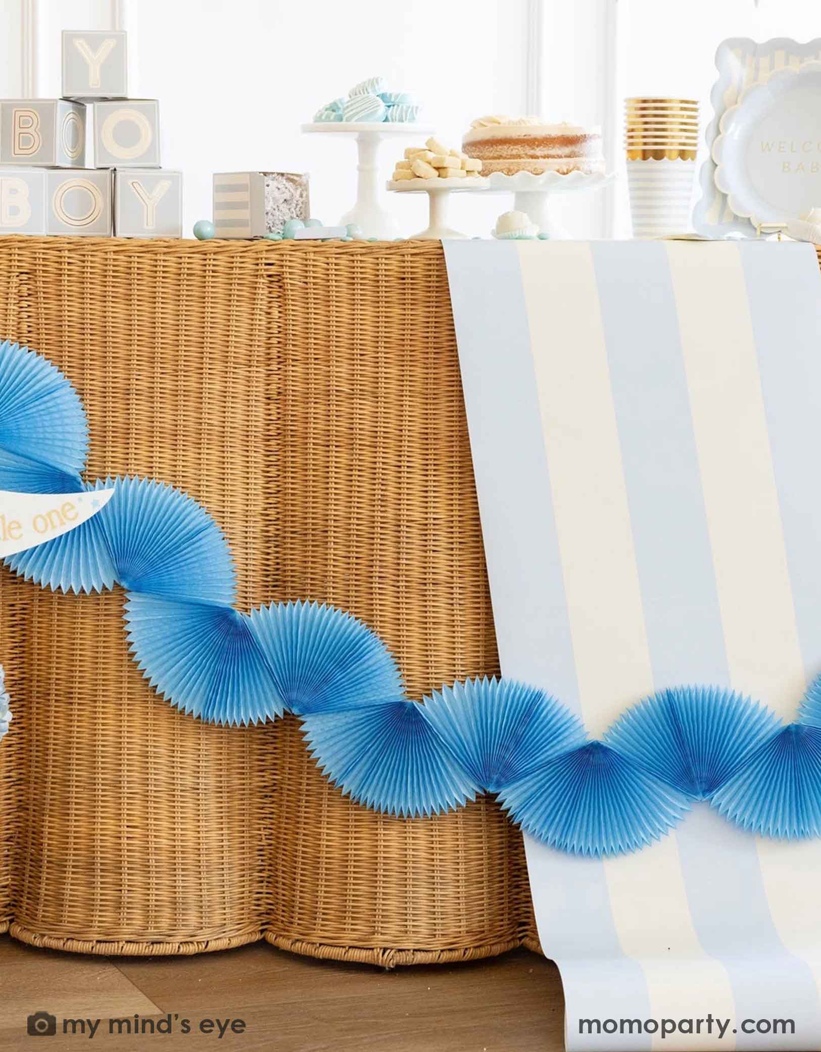 A modern and clean boy baby shower set up featuring straw-strung table covered with Momopartys's baby blue and white striped table runner. On the table there are various baby shower tablewares from Momopartys including party cups, treat boxes, plates and more. There's also a blue bunting banner hung across the table.