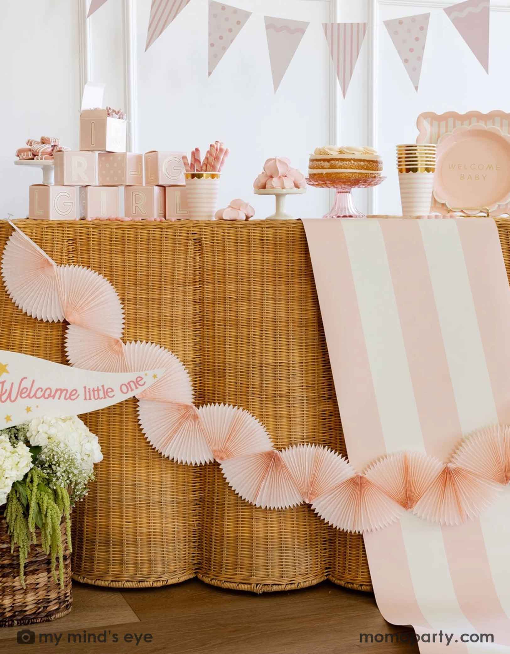 A modern and clean gril baby shower set up featuring straw-strung table covered with Momopartys's baby pink and white striped table runner. On the table there are various baby girl shower tablewares from Momopartys including party cups, treat boxes, plates and more. There's also a pastel pink bunting banner hung across the table and a "Welcome Little One" party pennant in the flower basket at the bottom.