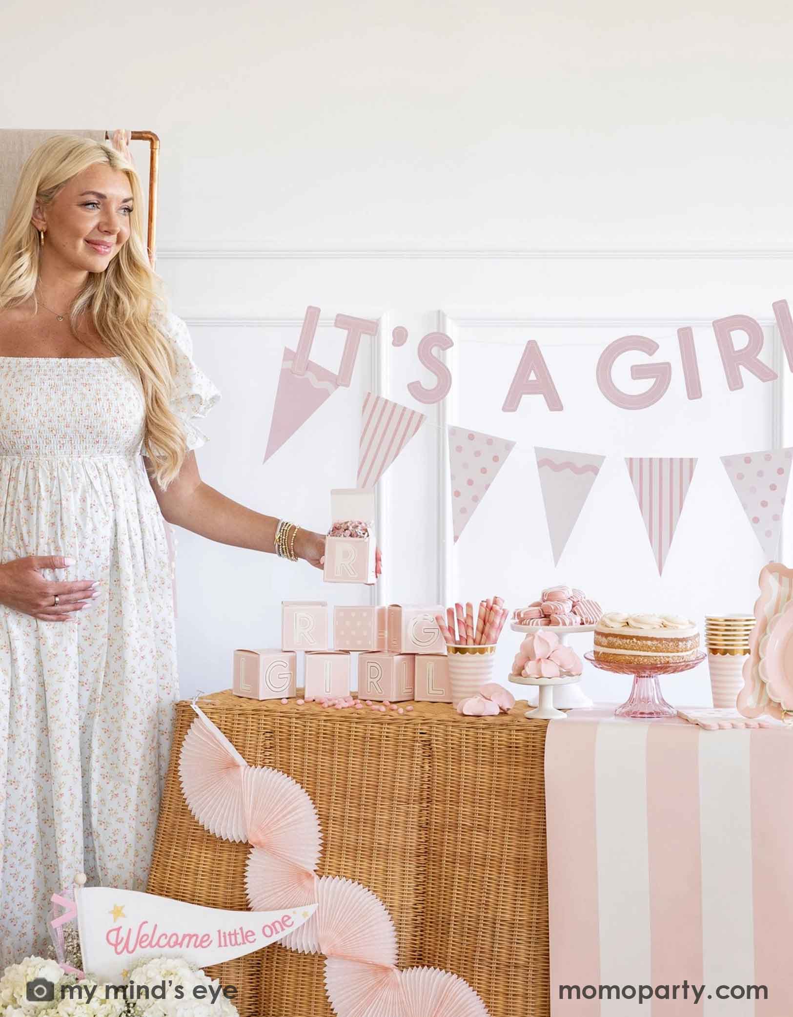 A pregnant woman standing next to a baby shower table with 'It's a Girl' banner and baby pink shower decorations and party supplies from Momopartys including tablewares, party banners, table runner and bunting garlands.