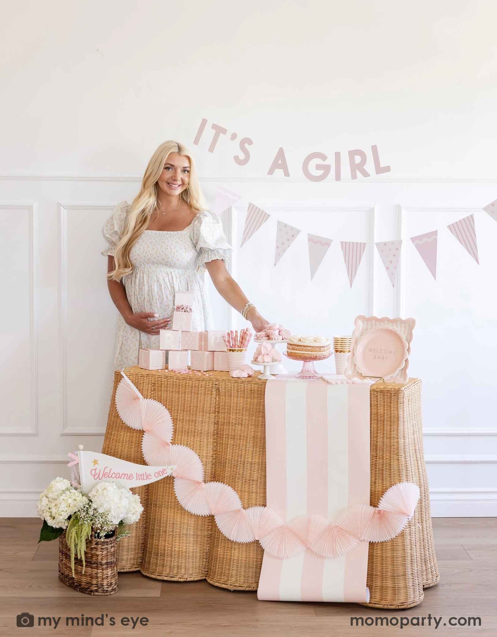 A pregnant woman standing next to a baby shower table with 'It's a Girl' banner and baby pink shower decorations and party supplies from Momopartys including tablewares, party banners, table runner and bunting garlands.