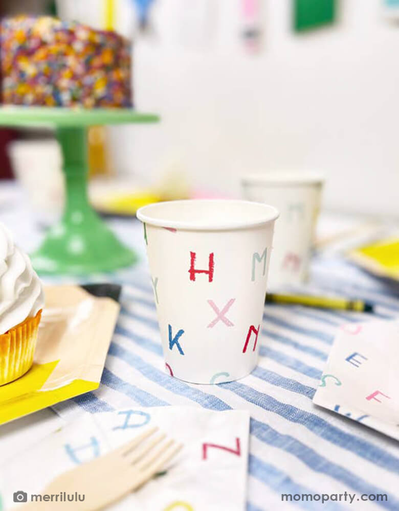 A close up shot of a back to school party table featuring Momopartys's alphabet letter patterned party cups by Merrilulu. In the back you can see Next to the tableware there's a cake topped with a pencil shaped cake topper with "Hello 2nd Grade". In the back there's a back to school party banner featuring colorful school themed pennants including crayon, glue bottle, notebook and more. All this makes a great inspo for a back to school party or first day of school celebration.
