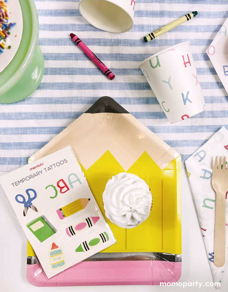 A kid's back to school party table filled with Momopartys's school themed party supplies by Merrilulu, including a pencil shaped plate, ABC letter patterned napkins, plates and party cups in bright fun colors, on top of the plate is a temporary tattoos sheet featuring colorful school themed illustrations. All this makes a great party inspo for kid's back to school party or first day of school celebration.