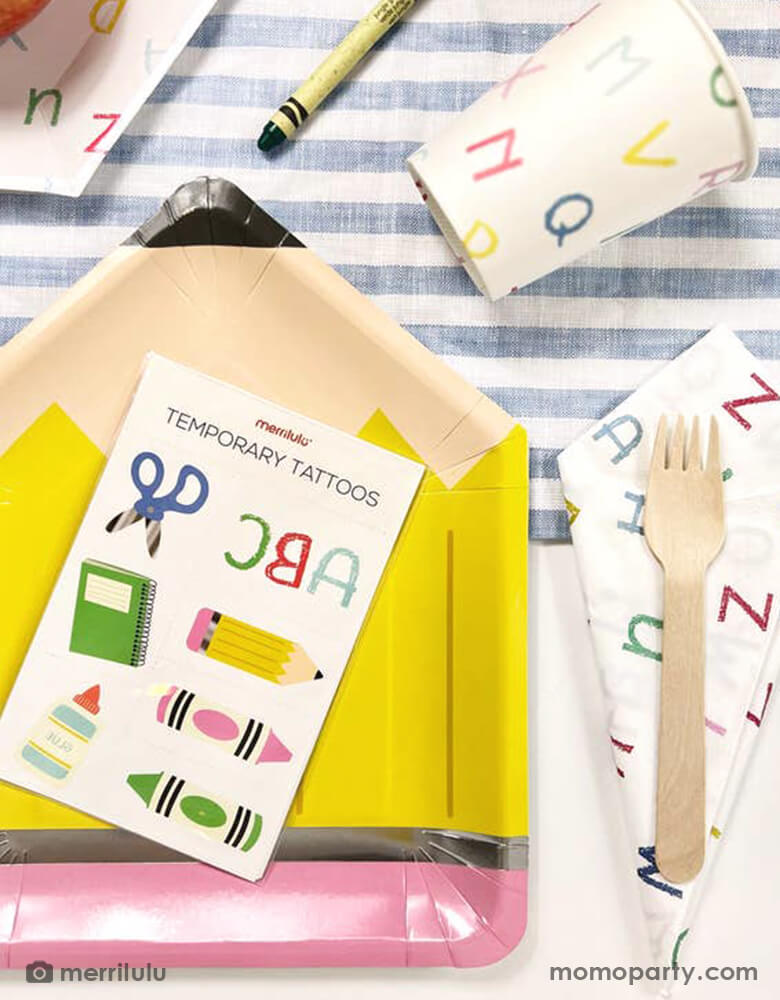 A back to school party table filled with Momopartys's school themed party supplies including a pencil shaped plate, ABC letter patterned napkins and party cups, on top of the plate is a temporary tattoos sheet featuring colorful school themed illustrations. All this makes a great party inspo for kid's back to school party or first day of school celebration.