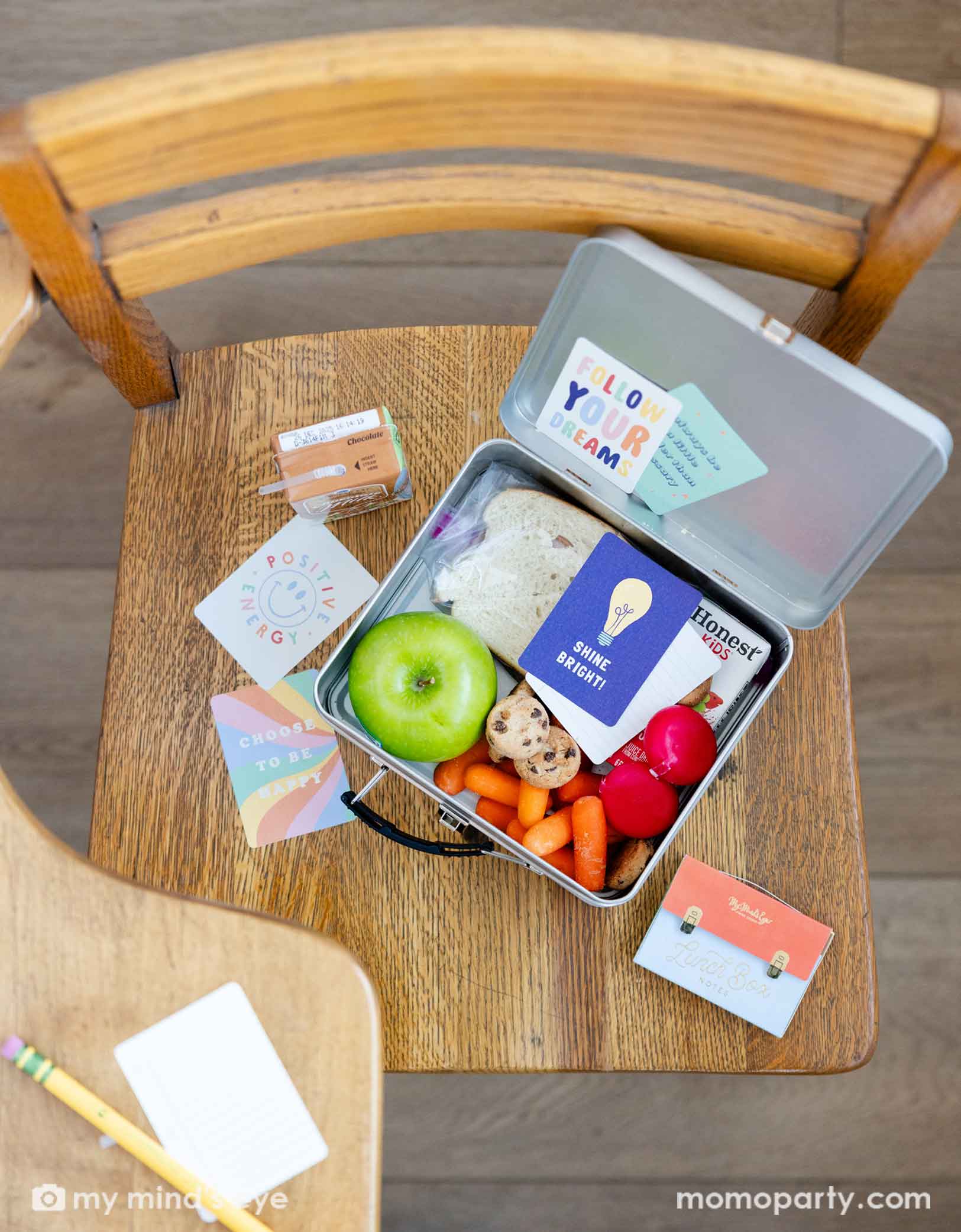 An overhead shot of a kid's lunch box on a school chair. In the lunch box includes a sandwich, an green apple, a juice box,   some cheese, veggies and cookies. There's a  card with "Shine Bright!" on it. Next to the lunch box are more lunchbox cards around and Momopartys's Lunch Box Cards package. 