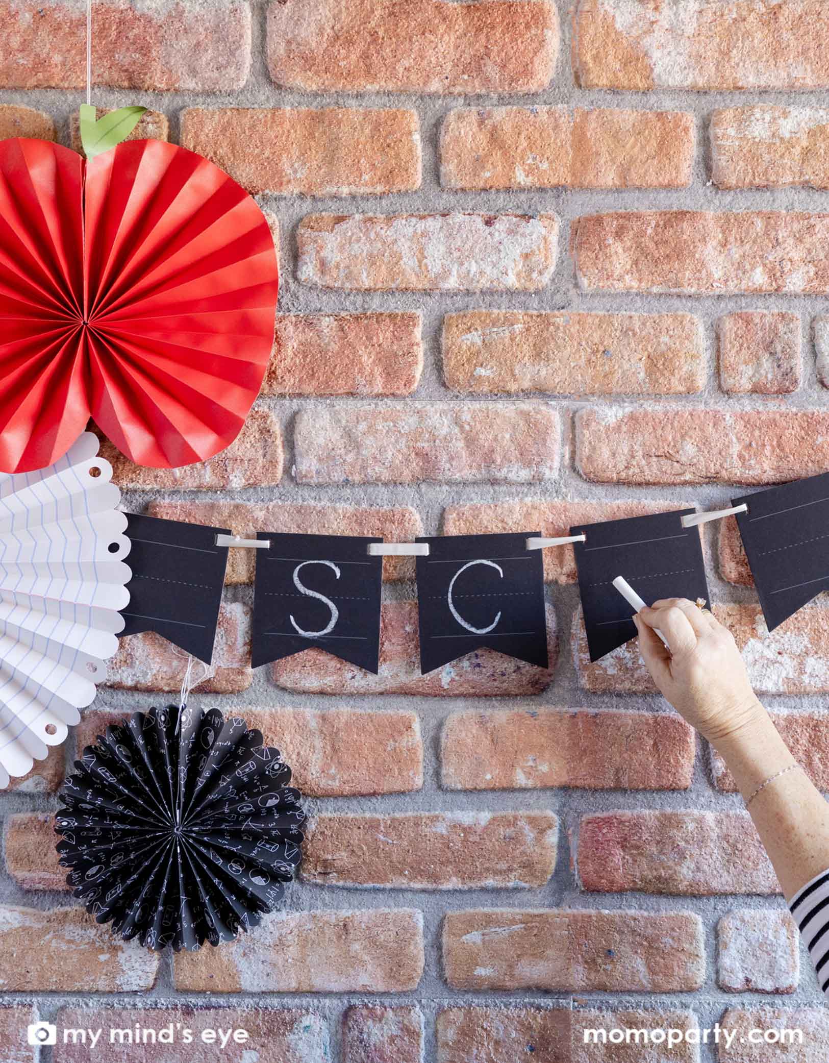 A brick wall decorated with Momopartys's back to school decorations including the Chalkboard Writable Paper Banner and the back to school party fans featuring an apple shaped fan and notebook patterned party fans. A hand is writing SC on the write-on party banner with a chalk. This banner lets you customize messages with chalk or chalk markers. Easy to write on and erase, it adds a fun, interactive touch to any celebration or learning space.