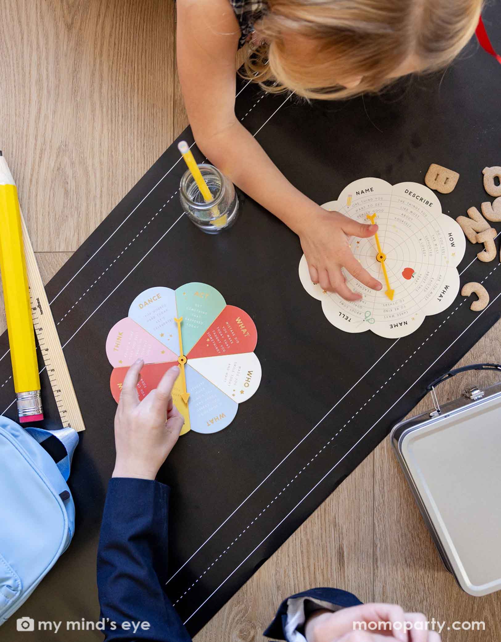 Two school-aged kids playing an activity spinning game on the floor covered with Momopartys's chalkboard paper table runner. On the runner there are jumbo school supplies including a ruler, a jumbo pencil, some alphabet cookies, a lunch box and its bag.